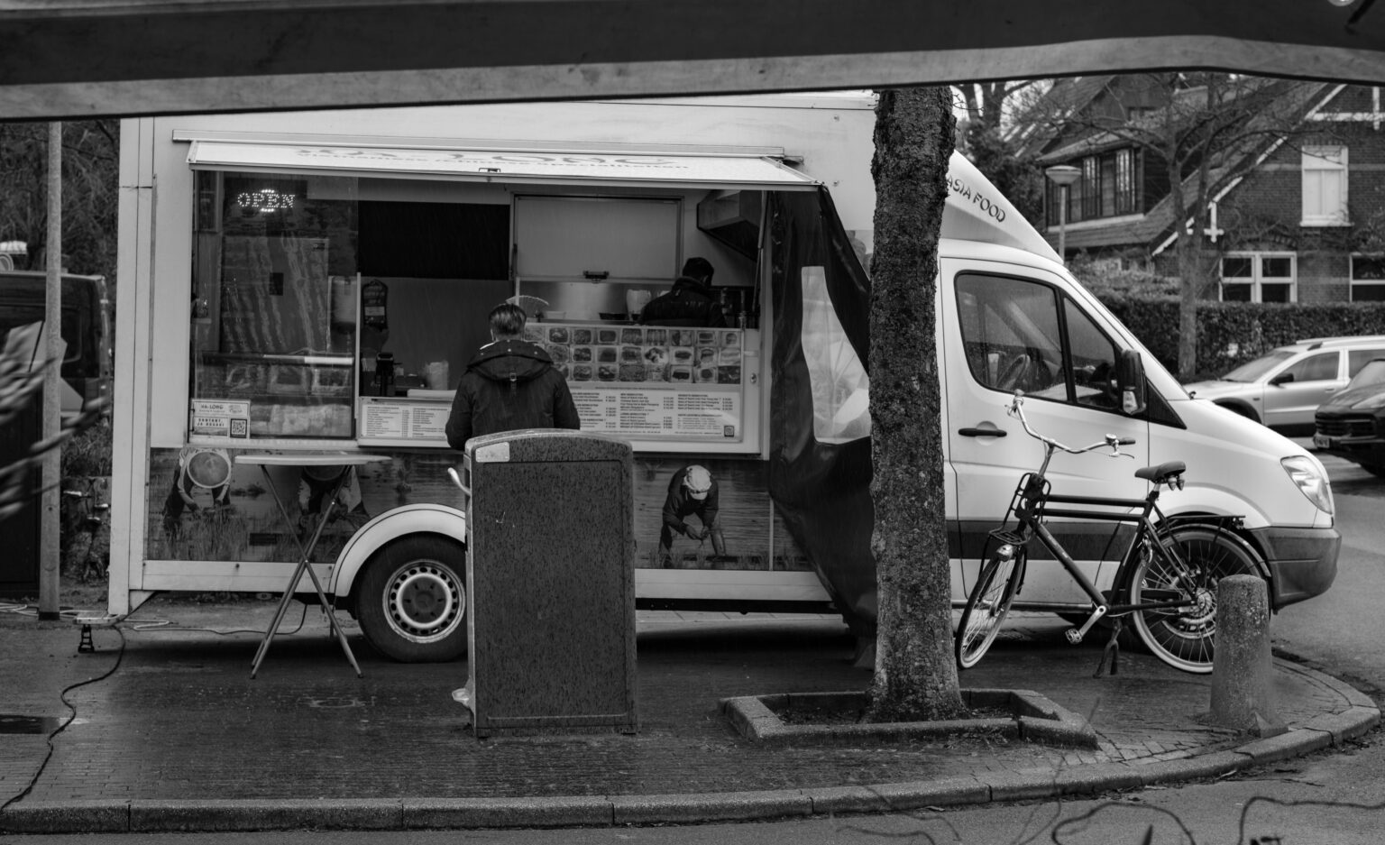 Black and white photo of a food truck with a man waiting, bicycle nearby, urban setting.