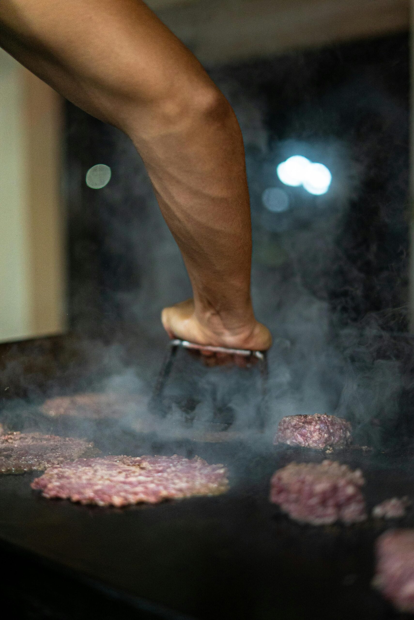 A chef's arm pressing burger patties on a steaming grill indoors, captured creatively.